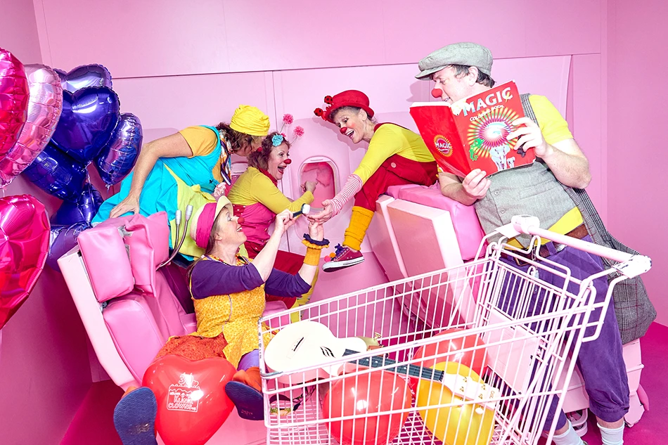 5 Clowns in bunten Kostümen interagieren fröhlich in einem pinken Raum mit Luftballons und Einkaufswagen