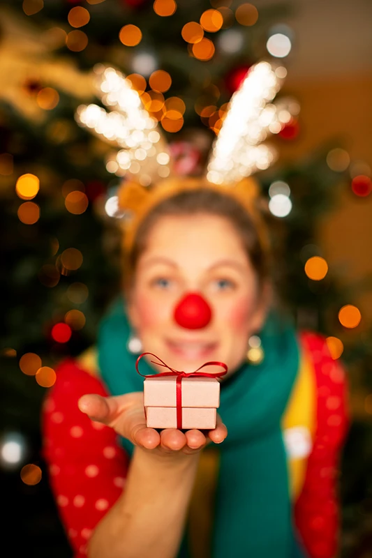 Klinikclownin präsentiert ein Geschenk vor geschmückten Weihnachtsbaum voller Lichter